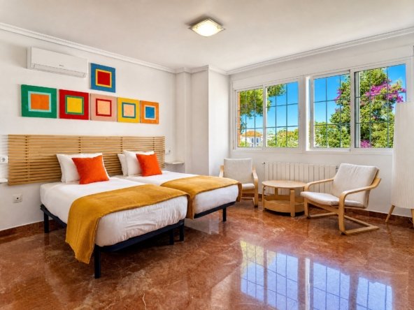 Dormitorio luminoso con dos camas, cuadros de colores, zona de estar y ventanas con vistas a pinos y buganvilla