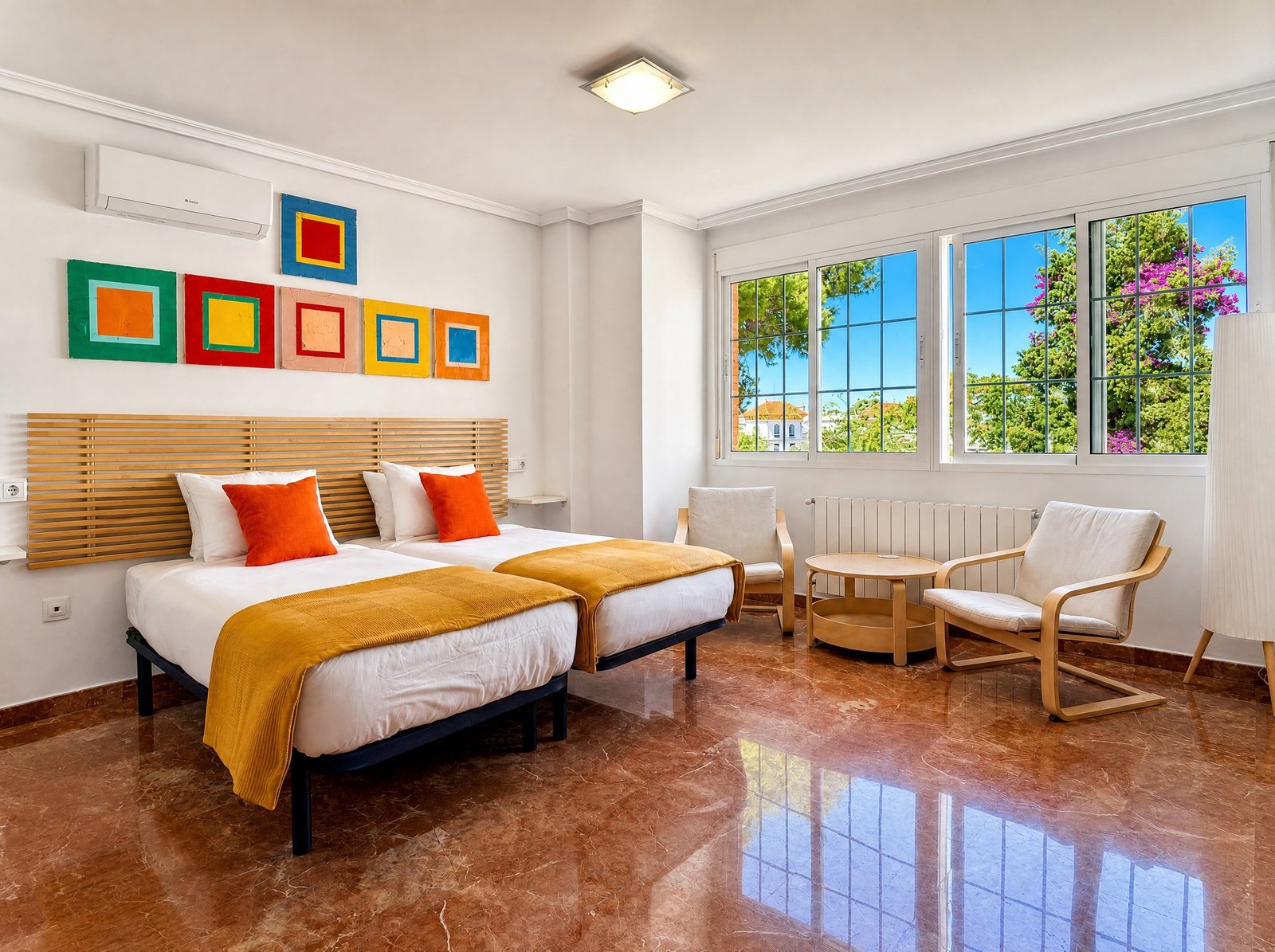 Dormitorio luminoso con dos camas, cuadros de colores, zona de estar y ventanas con vistas a pinos y buganvilla